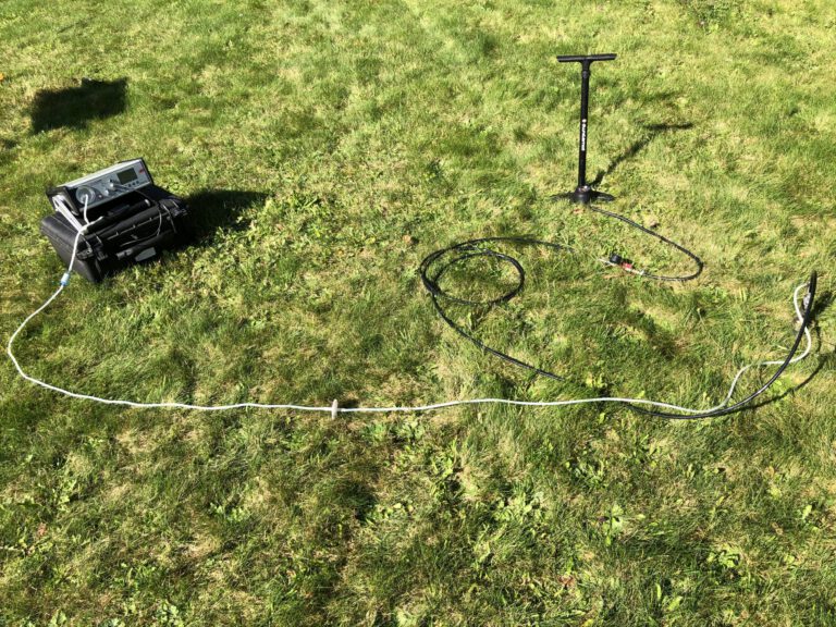 Das Bild zeigt eine Messapparatur auf einer grünen Wiese. Links steht ein schwarzer Transportkoffer mit einem silbernen Messgerät darauf, von dem ein langes weißes Kabel über den Boden verläuft. Rechts ist eine schwarze Handpumpe und weitere Kabel zu sehen, die für die Messung im Freien genutzt werden.