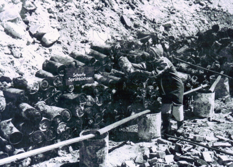 Das Bild zeigt eine große Menge von länglichen Metallobjekten, die in mehreren Reihen an einem Erdhang gestapelt sind. Die Objekte wirken stark abgenutzt und liegen teilweise lose aufeinander. Im Vordergrund sind einige Behälter und Stangen zu sehen, die vermutlich zur Sicherung oder Handhabung dienen. Die Szene deutet auf eine Arbeitsumgebung im Freien hin, in der mit alten militärischen Materialien oder Munition umgegangen wird.