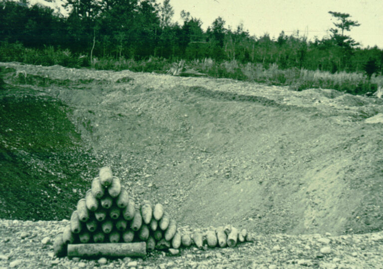 Das Bild zeigt eine große, offene Grube in einem Erdgelände, umgeben von Bäumen im Hintergrund. Im Vordergrund ist ein Stapel von länglichen Metallobjekten zu sehen, die pyramidenförmig angeordnet sind, sowie einige weitere Objekte daneben auf dem Boden. Die Szene wirkt wie ein ehemaliges militärisches Gelände oder eine Ablagestelle für Munition.