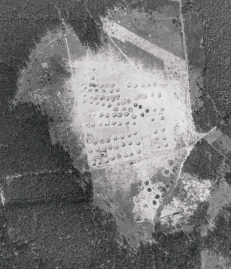 Das Bild zeigt eine Luftaufnahme eines großen, offenen Geländes, das von Waldflächen umgeben ist. In der Mitte des Geländes sind zahlreiche kreisförmige Vertiefungen im Boden zu erkennen, die in einem regelmäßigen Muster angeordnet sind. Die Umgebung wirkt stark verändert und weist Spuren von Erdbewegungen oder Explosionen auf, was auf eine ehemalige militärische Nutzung oder ein Testgelände hindeutet.