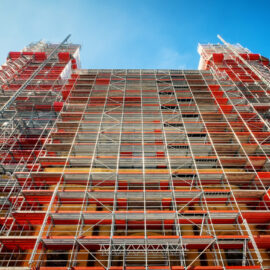 Ein hohes Gebäude ist vollständig von einem Gerüst umgeben, das aus zahlreichen Metallstreben und roten sowie silbernen Plattformen besteht. Die Konstruktion wirkt sehr komplex und reicht bis in den klaren blauen Himmel, was auf umfangreiche Bau- oder Sanierungsarbeiten hinweist. Die Perspektive zeigt das Gerüst von unten nach oben, wodurch die Höhe und Stabilität der Struktur besonders deutlich wird.