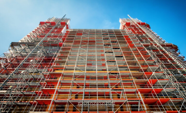 Ein hohes Gebäude ist vollständig von einem Gerüst umgeben, das aus zahlreichen Metallstreben und roten sowie silbernen Plattformen besteht. Die Konstruktion wirkt sehr komplex und reicht bis in den klaren blauen Himmel, was auf umfangreiche Bau- oder Sanierungsarbeiten hinweist. Die Perspektive zeigt das Gerüst von unten nach oben, wodurch die Höhe und Stabilität der Struktur besonders deutlich wird.