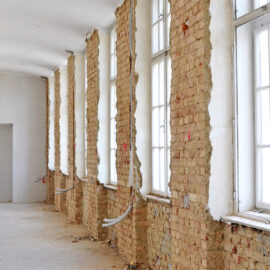 Das Bild zeigt einen langen Flur mit hohen Fenstern auf der rechten Seite, deren Rahmen weiß gestrichen sind. Die Wand unter den Fenstern besteht aus freigelegtem Mauerwerk, während die gegenüberliegende Wand bereits verputzt und weiß gestrichen ist. Auf dem Boden liegen einige Kabel, und die gesamte Szene vermittelt den Eindruck von Renovierungs- oder Sanierungsarbeiten im Innenbereich.