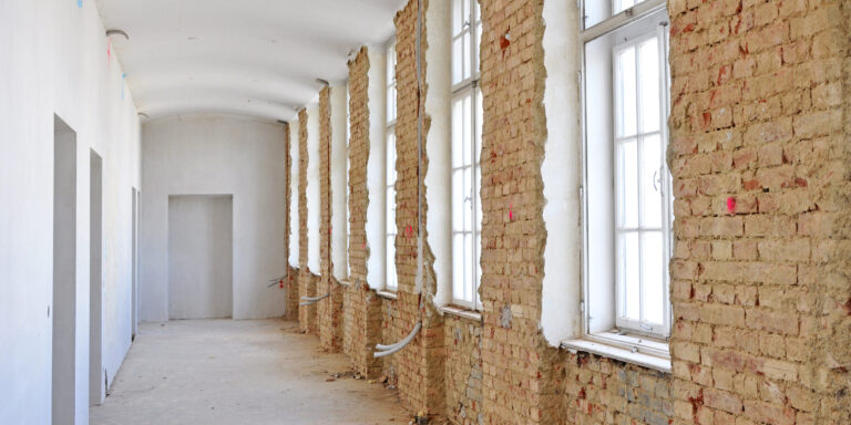 Das Bild zeigt einen langen Flur mit hohen Fenstern auf der rechten Seite, deren Rahmen weiß gestrichen sind. Die Wand unter den Fenstern besteht aus freigelegtem Mauerwerk, während die gegenüberliegende Wand bereits verputzt und weiß gestrichen ist. Auf dem Boden liegen einige Kabel, und die gesamte Szene vermittelt den Eindruck von Renovierungs- oder Sanierungsarbeiten im Innenbereich.
