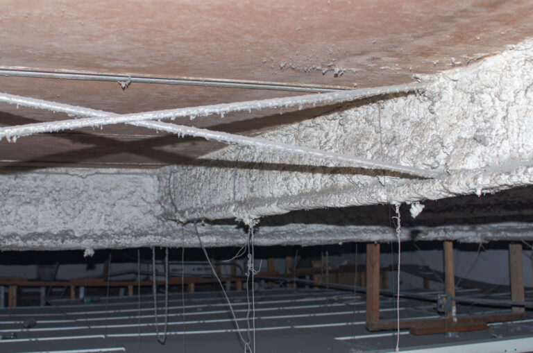 Das Bild zeigt eine Betondecke mit einer dicken, weißen Beschichtung, die unregelmäßig aufgetragen ist und teilweise auf die Metallstreben überläuft. Mehrere dünne Drähte hängen von der Decke herab, vermutlich zur Befestigung von abgehängten Elementen.