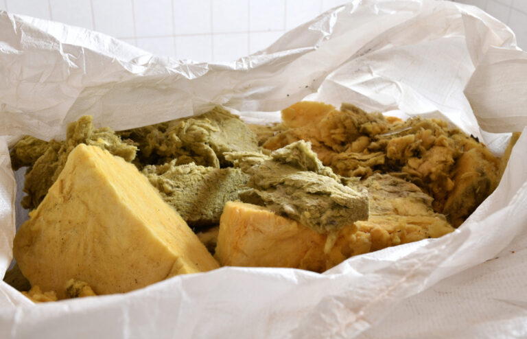 Das Bild zeigt eine weiße Kunststofftüte, die mit großen Stücken von gelblicher und grünlicher Mineralwolle gefüllt ist. Die Dämmmaterialien wirken stark gebraucht und teilweise zerfasert, was auf eine Entfernung aus einem Gebäude hindeutet. Im Hintergrund ist eine helle, geflieste Fläche zu erkennen, die den Eindruck einer Innenaufnahme vermittelt.