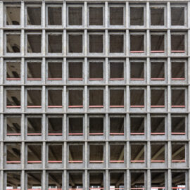 Das Bild zeigt die Fassade eines Rohbaus mit einer regelmäßigen Gitterstruktur aus Beton. Die quadratischen Öffnungen sind gleichmäßig angeordnet und geben den Blick auf die leeren Etagen des Gebäudes frei. Zwischen den Betonrahmen verlaufen horizontale rote Linien, die vermutlich als Sicherheitsmarkierungen dienen.