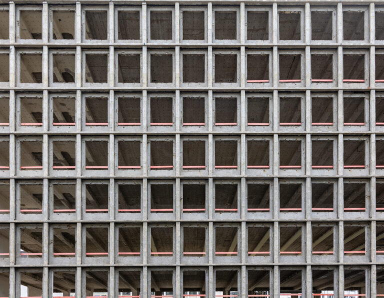Das Bild zeigt die Fassade eines Rohbaus mit einer regelmäßigen Gitterstruktur aus Beton. Die quadratischen Öffnungen sind gleichmäßig angeordnet und geben den Blick auf die leeren Etagen des Gebäudes frei. Zwischen den Betonrahmen verlaufen horizontale rote Linien, die vermutlich als Sicherheitsmarkierungen dienen.