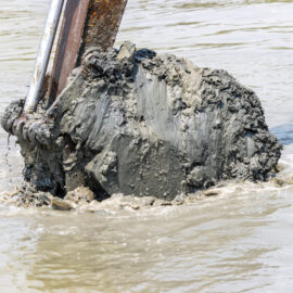Das Bild zeigt eine große Baggerschaufel, die aus dem Wasser gehoben wird und mit dunklem, nassem Schlamm bedeckt ist. Um die Schaufel herum ist trübes Wasser zu sehen, das durch die Bewegung aufgewühlt wirkt. Im Hintergrund sind Teile weiterer metallischer Strukturen erkennbar, die auf eine Arbeitsumgebung im Wasser hinweisen.