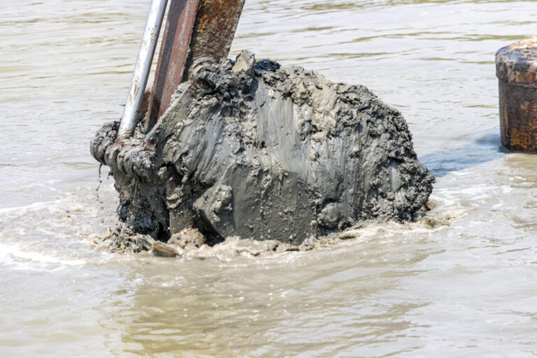 Das Bild zeigt eine große Baggerschaufel, die aus dem Wasser gehoben wird und mit dunklem, nassem Schlamm bedeckt ist. Um die Schaufel herum ist trübes Wasser zu sehen, das durch die Bewegung aufgewühlt wirkt. Im Hintergrund sind Teile weiterer metallischer Strukturen erkennbar, die auf eine Arbeitsumgebung im Wasser hinweisen.