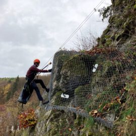 Das Bild zeigt eine Person, die mit Kletterausrüstung an einer steilen Felswand arbeitet. Die Felsoberfläche ist teilweise mit Vegetation und einem engmaschigen Drahtnetz bedeckt, das zur Sicherung angebracht wurde. Die Person ist mit Seilen gesichert und trägt einen Helm, während im Hintergrund eine bewaldete Landschaft und einige Gebäude zu erkennen sind.