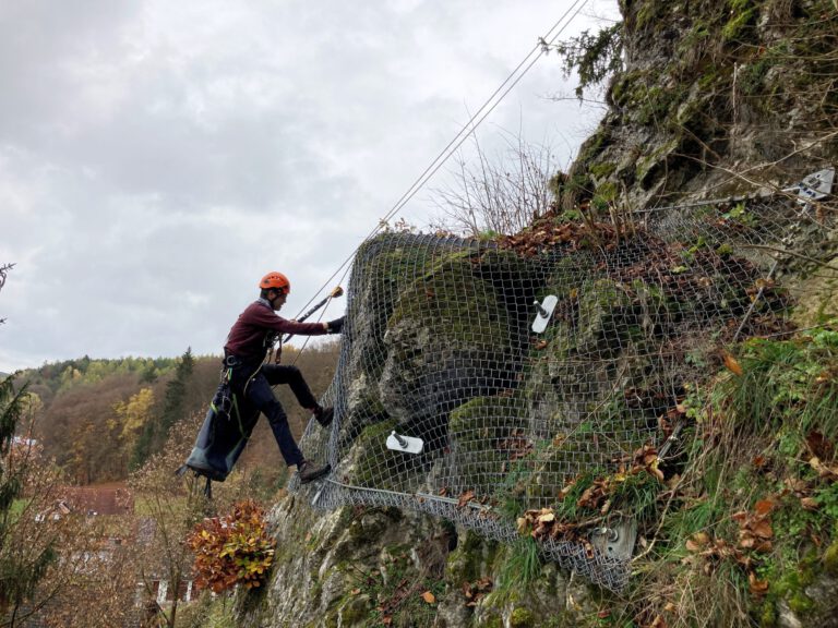 Das Bild zeigt eine Person, die mit Kletterausrüstung an einer steilen Felswand arbeitet. Die Felsoberfläche ist teilweise mit Vegetation und einem engmaschigen Drahtnetz bedeckt, das zur Sicherung angebracht wurde. Die Person ist mit Seilen gesichert und trägt einen Helm, während im Hintergrund eine bewaldete Landschaft und einige Gebäude zu erkennen sind.