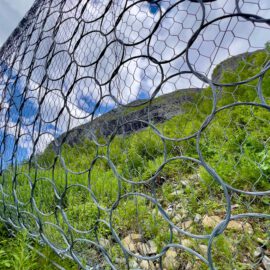 Das Bild zeigt ein Steinschlagschutznetz, das aus großen, kreisförmigen Metallringen besteht und an einer Böschung installiert ist. Hinter dem Netz ist eine grüne Hangfläche mit Gras und vereinzelten Steinen zu sehen, die zu einem felsigen Berghang ansteigt. Im Hintergrund erstreckt sich ein blauer Himmel mit weißen Wolken, was der Szene einen offenen und natürlichen Charakter verleiht.