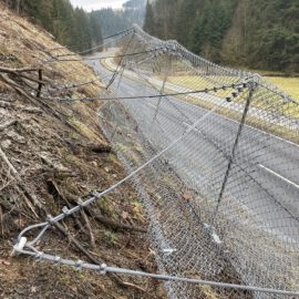 Das Bild zeigt ein Steinschlagschutznetz, das entlang einer Böschung oberhalb einer Straße installiert ist. Das Netz besteht aus engmaschigem Drahtgeflecht, das mit mehreren Stahlseilen und Befestigungselementen am Hang verankert ist. Im Hintergrund verläuft eine asphaltierte Straße durch eine offene Landschaft mit Wiesen und dichtem Wald, während die Böschung im Vordergrund von Erde, Wurzeln und vereinzelten Ästen bedeckt ist.