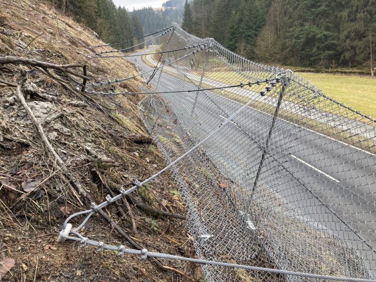 Das Bild zeigt ein Steinschlagschutznetz, das entlang einer Böschung oberhalb einer Straße installiert ist. Das Netz besteht aus engmaschigem Drahtgeflecht, das mit mehreren Stahlseilen und Befestigungselementen am Hang verankert ist. Im Hintergrund verläuft eine asphaltierte Straße durch eine offene Landschaft mit Wiesen und dichtem Wald, während die Böschung im Vordergrund von Erde, Wurzeln und vereinzelten Ästen bedeckt ist.