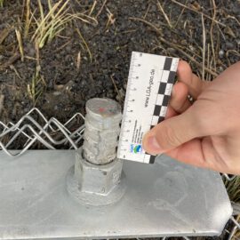Das Bild zeigt eine Nahaufnahme eines metallischen Befestigungselements, das auf einer flachen Platte montiert ist. An dem Element ist ein Drahtseil befestigt, und daneben hält eine Hand einen Maßstab, um die Größe des Bauteils zu dokumentieren. Im Hintergrund liegt dunkle Erde mit einigen Grashalmen, und Teile eines Maschendrahtzauns sind ebenfalls sichtbar.