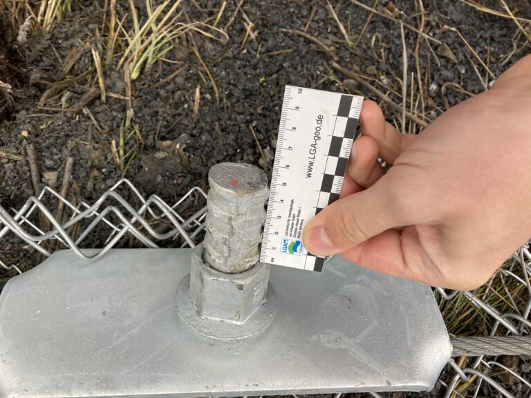 Das Bild zeigt eine Nahaufnahme eines metallischen Befestigungselements, das auf einer flachen Platte montiert ist. An dem Element ist ein Drahtseil befestigt, und daneben hält eine Hand einen Maßstab, um die Größe des Bauteils zu dokumentieren. Im Hintergrund liegt dunkle Erde mit einigen Grashalmen, und Teile eines Maschendrahtzauns sind ebenfalls sichtbar.
