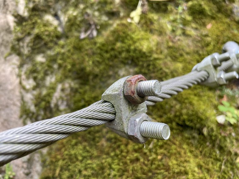 Das Bild zeigt ein dickes Stahlseil, das mit einer Metallklemme und zwei Schrauben befestigt ist. Die Klemme hält mehrere verdrillte Drahtstränge zusammen und sorgt für Stabilität. Im Hintergrund ist eine mit Moos bedeckte Felsoberfläche zu sehen, die eine natürliche Umgebung andeutet.