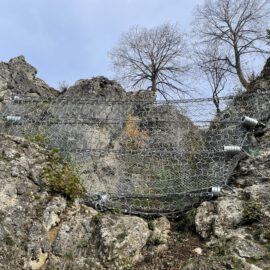 Das Bild zeigt eine steile Felswand, die mit mehreren stabilen Drahtnetzen gesichert ist. Die Netze sind mit großen Federn und Befestigungselementen an den Felsen verankert, um Steinschläge zu verhindern. Im oberen Bereich stehen einige kahle Bäume vor einem hellen Himmel.