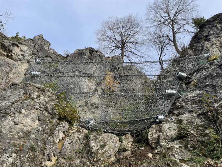 Das Bild zeigt eine steile Felswand, die mit mehreren stabilen Drahtnetzen gesichert ist. Die Netze sind mit großen Federn und Befestigungselementen an den Felsen verankert, um Steinschläge zu verhindern. Im oberen Bereich stehen einige kahle Bäume vor einem hellen Himmel.