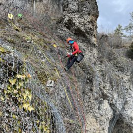 Eine Person arbeitet an einer steilen Felswand und ist mit Kletterausrüstung sowie Sicherheitsseilen gesichert. Die Felswand ist teilweise mit Drahtnetzen bedeckt, die vermutlich zur Sicherung gegen Steinschlag dienen. Im Hintergrund sind einige Bäume und ein bewölkter Himmel zu sehen.